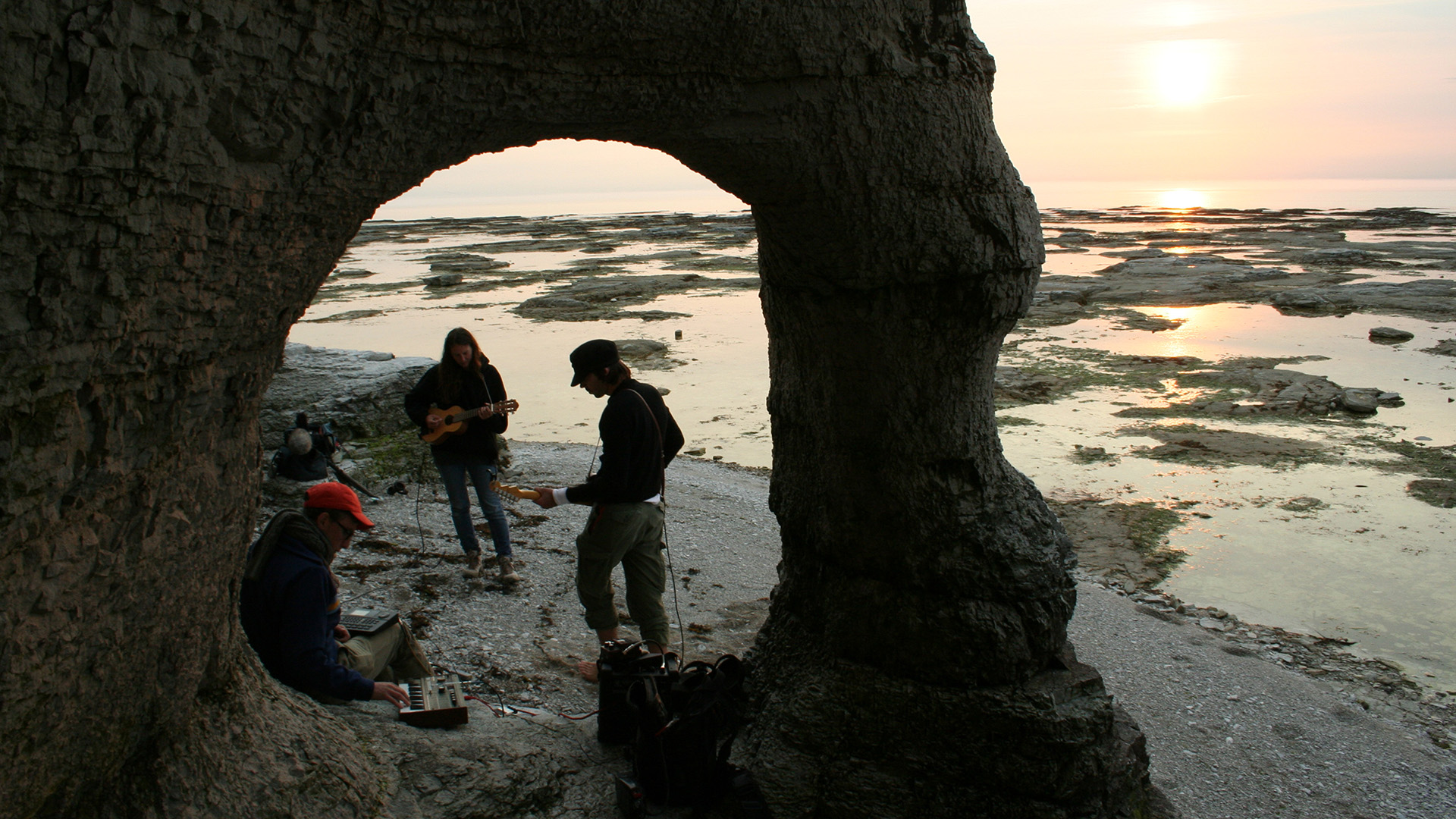 Musicians perform beneath a natural rock arch on a rocky shoreline at sunset, recording a session for the film.