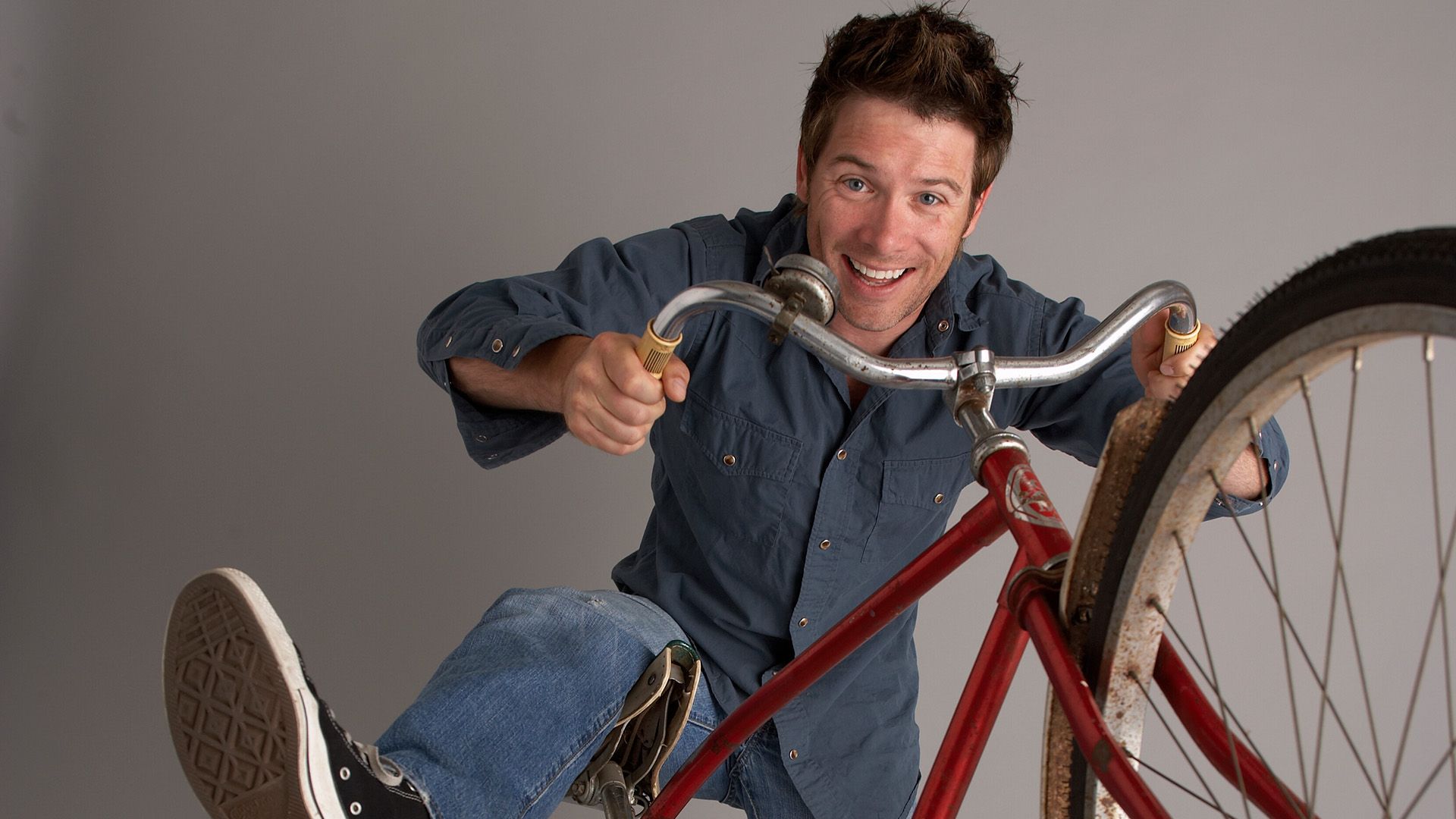 Host Jeff Douglas grins while perched on an old red bicycle, gripping the handlebars.