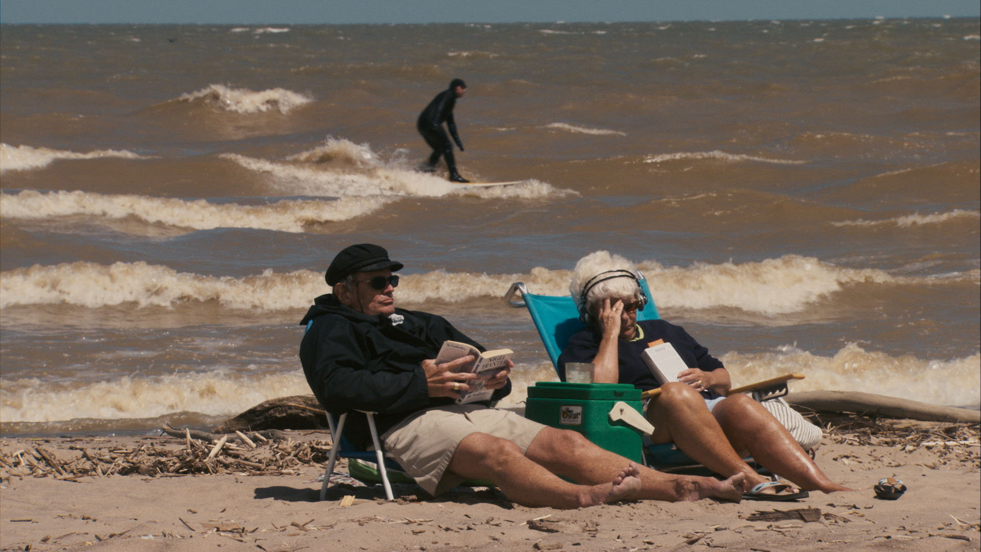Two people relax in beach chairs reading, while a surfer rides a wave on a Great Lake behind them.