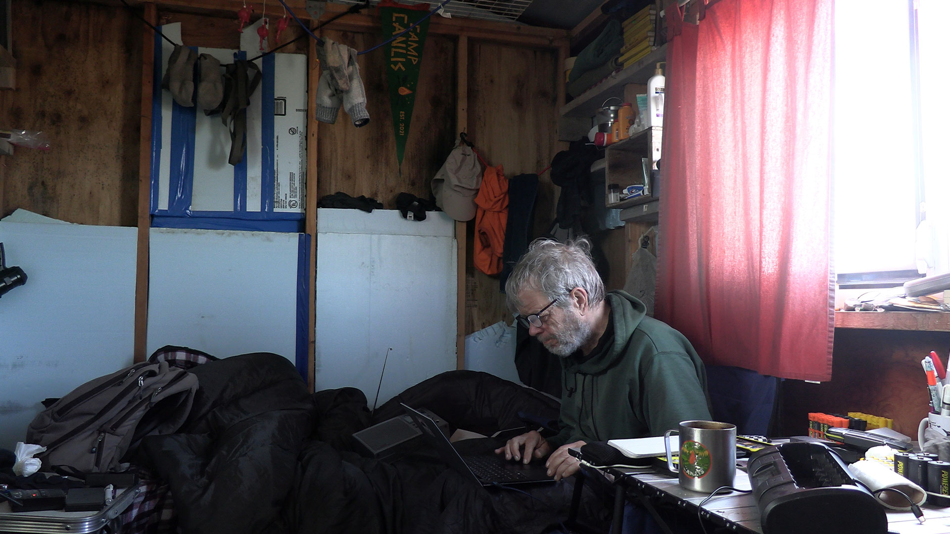 George Divoky works at a laptop inside his Arctic field camp, surrounded by expedition gear and a 'Camp Canlis' pennant.