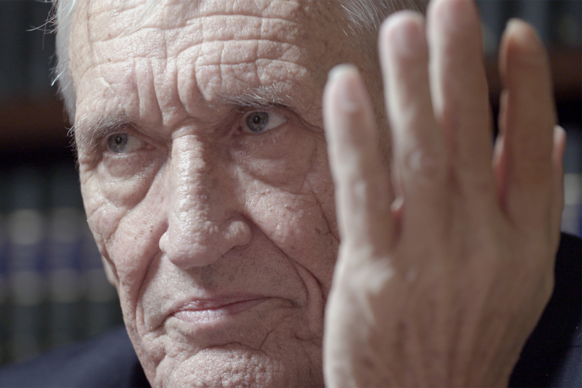 Close-up of an elderly man's weathered face, hand raised, mid-conversation.