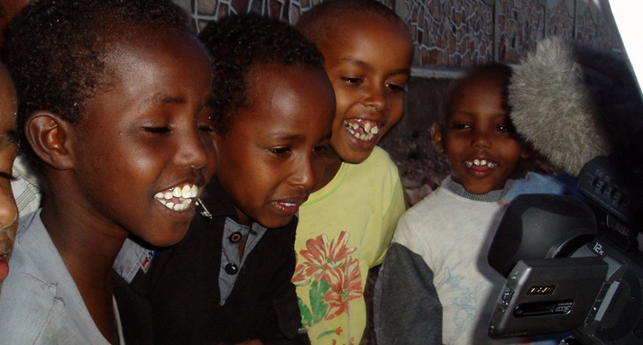 A group of children smile and crowd around a video camera, watching themselves on the viewfinder.