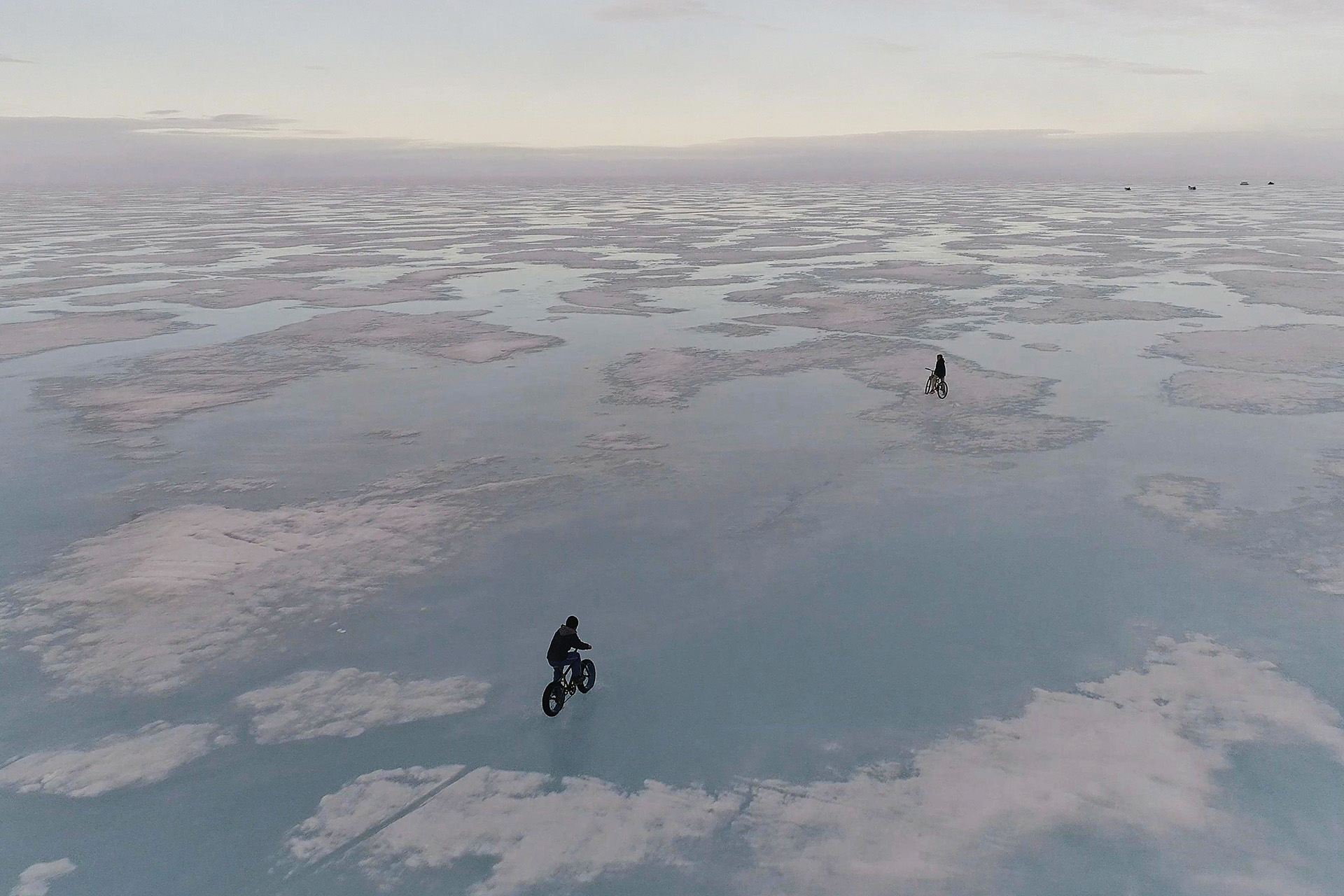 Two cyclists ride across a vast, reflective expanse of melting sea ice at dusk.