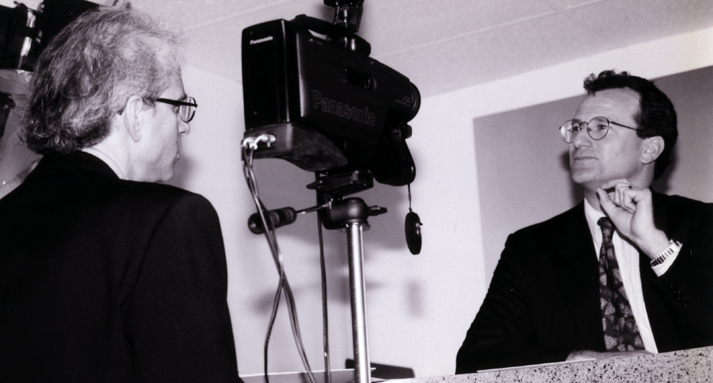 A man is interviewed on camera by an off-screen host, black-and-white production still.