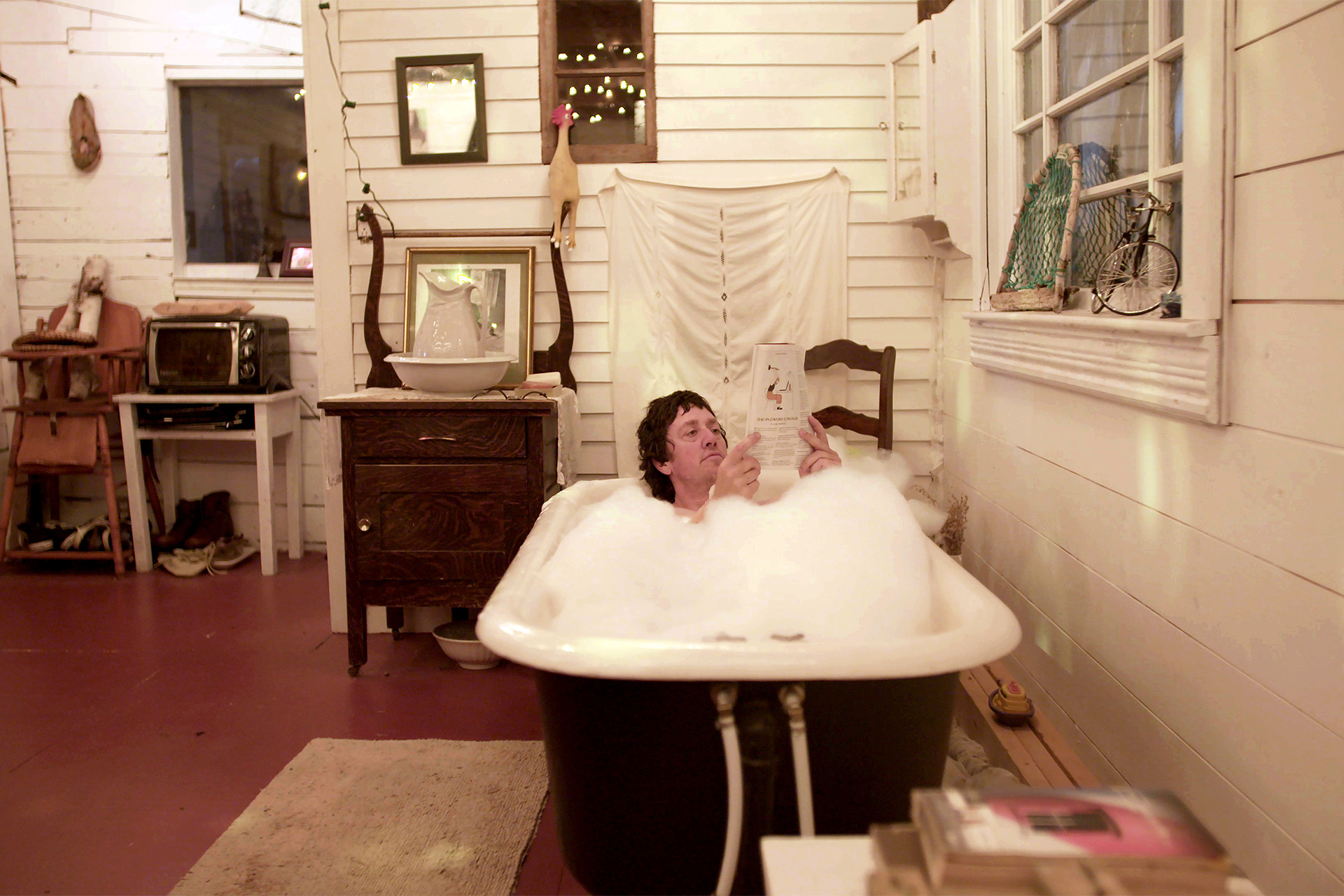 A man relaxes in a claw-foot bathtub filled with bubbles, reading a magazine in a rustic, plant-and-antique-filled room.